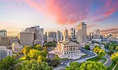 Aerial view of downtown Nashville, Tennessee, at sunset with vibrant pink clouds above the skyline. The Tennessee State Capitol stands prominently in the foreground, surrounded by green trees and curving roads.