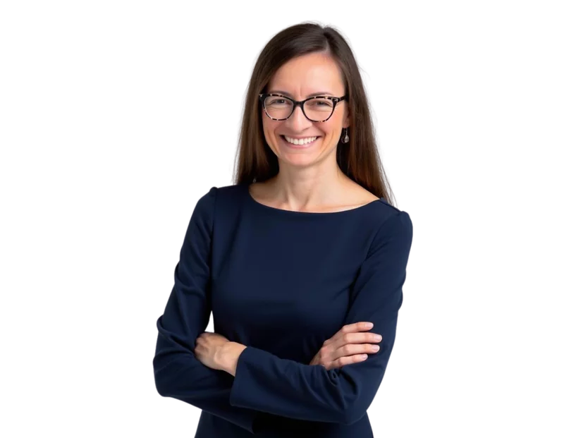 Smiling woman with long brown hair and glasses wearing a navy blue dress. She is standing with arms crossed against a transparent background.