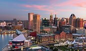 Aerial view of Baltimore’s Inner Harbor at sunset, with waterfront buildings reflecting warm pink and orange hues. The skyline features a mix of historic brick structures and modern high-rises, with the harbor waters in the foreground.