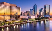 Evening view of downtown Jacksonville, Florida, with glass skyscrapers reflecting a vivid sunset. The St. Johns River runs along the foreground, its calm waters mirroring the city lights and colorful sky.