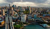 Aerial view of downtown Brooklyn, New York, with the Manhattan Bridge extending across the East River in the foreground. The skyline features a mix of modern high-rises and historic brick buildings surrounded by green spaces.