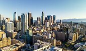 A bright daytime view of a modern city skyline with tall glass and steel skyscrapers rising above dense mid-rise buildings. Mountains are visible in the background under a clear blue sky.