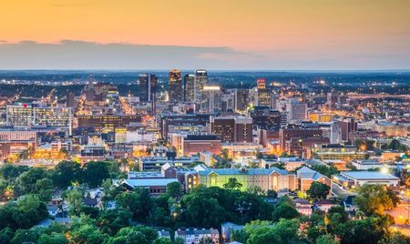 A panoramic view of a city skyline at dusk, with downtown high-rise buildings glowing under warm sunset light. The foreground shows tree-lined neighborhoods transitioning into the brightly lit urban core.