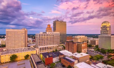 Downtown Winston-Salem, North Carolina, at sunset with a colorful sky casting warm light on its mix of historic and modern buildings. The distinctive rounded dome of the Winston Tower stands out among the skyline.