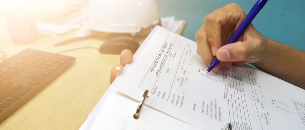 Close up of an engineer holding a clipboard and writing on a preventive maintenance checklist with a blue pen. A hard hat, computer keyboard, and desk equipment are visible in the background, suggesting a technical workspace.