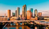Downtown Tampa, Florida, at golden hour with sunlight reflecting off its tall glass and brick skyscrapers. A bridge crosses the Hillsborough River in the foreground, connecting the urban core with surrounding neighborhoods.