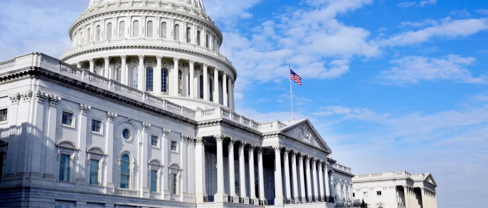 Wide angle view of the United States Capitol Building in Washington DC, featuring the iconic white dome and neoclassical columns under a bright blue sky. The American flag flies above the façade, emphasizing the building’s role as a public government land