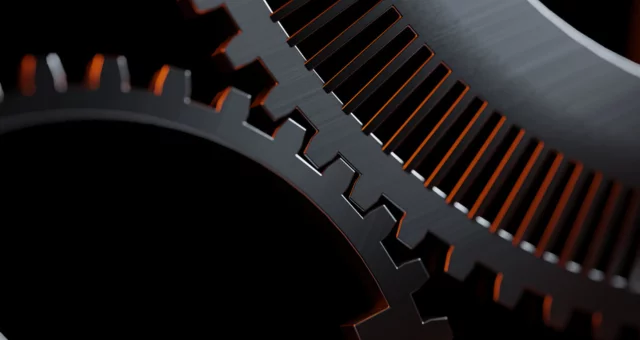 Close-up view of two interlocking metallic gears with sharp, polished teeth. The edges are highlighted by a warm orange light, contrasting with the dark, shadowed background.