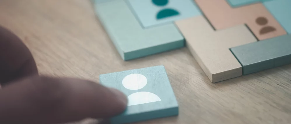 Close up of a hand placing a light colored geometric tangram piece onto a wooden puzzle board. Several pastel shapes featuring small people icons are arranged into a larger square pattern, showing the tangram puzzle in progress. The image captures the mom