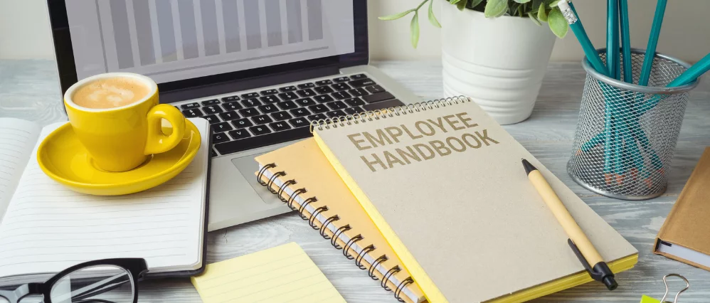 Employee handbook notebook placed on an office desk beside a laptop, yellow coffee cup, and stationery items. The spiral bound cover clearly reads Employee Handbook, with a pen resting on top.