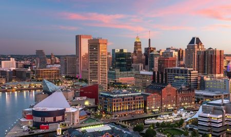 Aerial view of Baltimore’s Inner Harbor at sunset, with waterfront buildings reflecting warm pink and orange hues. The skyline features a mix of historic brick structures and modern high-rises, with the harbor waters in the foreground.