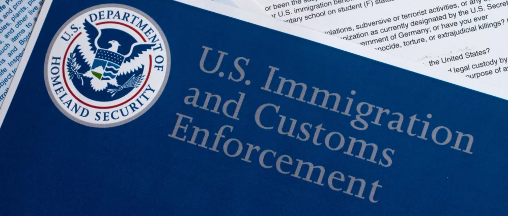 Close up of a blue document labeled U.S. Immigration and Customs Enforcement with the seal of the U.S. Department of Homeland Security visible at the top. Official paperwork with printed text appears partially underneath the cover.