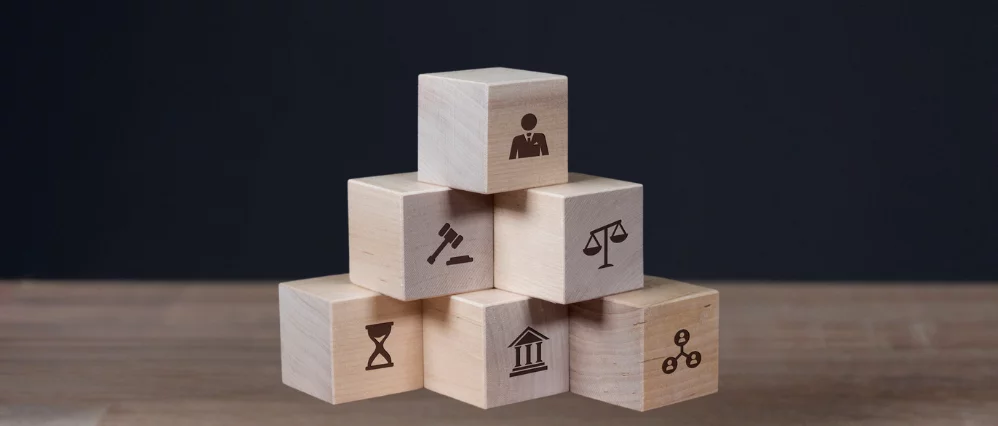 Pyramid stack of wooden cube blocks arranged on a table against a dark blue background, each cube printed with a black icon. The icons include a businessperson, a gavel, scales of justice, an hourglass, a courthouse building, and a connected network symbo