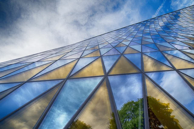 A modern glass building with triangular window panels reflects a vivid blue sky, white clouds, and green trees. The low-angle perspective emphasizes the geometric pattern as the structure appears to stretch infinitely upward.