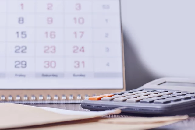 A desk scene shows a blurred monthly calendar in the background with a gray calculator and manila envelope in focus on a wooden surface. The setup suggests planning, scheduling, or financial calculations.