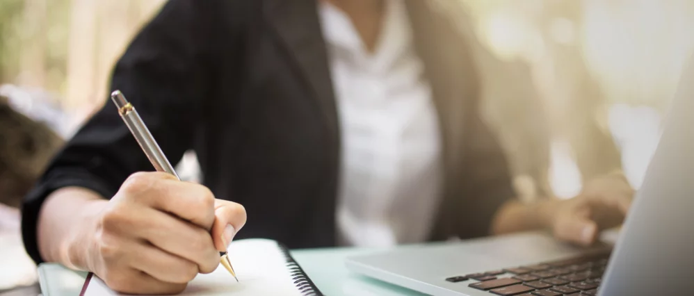 Close up of a person in business attire writing in a notebook while using a laptop at a desk. Warm sunlight filters across the scene, creating a focused and professional atmosphere.