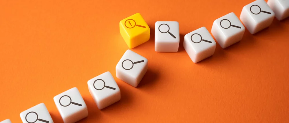 Diagonal row of white cube blocks with magnifying glass icons arranged on a bright orange background, with one yellow cube standing out among them. The yellow block features a magnifying glass with an exclamation mark, drawing attention to a specific issu