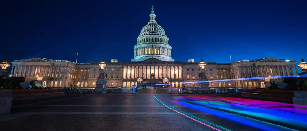 Wide angle view of the United States Capitol building illuminated at night under a deep blue sky. The white dome and neoclassical façade glow with warm lights, while colorful light trails streak across the foreground walkway.