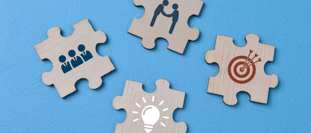 Overhead view of wooden jigsaw puzzle pieces placed on a blue background, each featuring business themed icons. Symbols include a team group, handshake, target with arrows, and a light bulb for ideas.