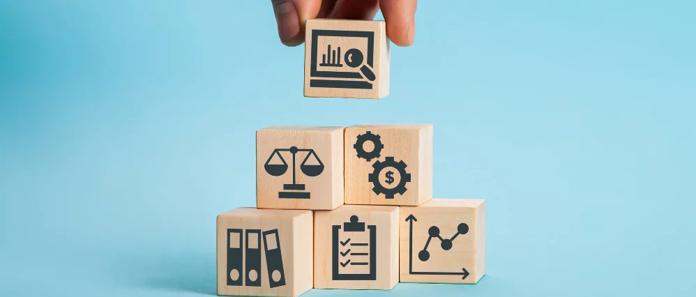 Close up of a hand placing a wooden cube with a chart and magnifying glass icon on top of a stack of business themed blocks. The cubes display symbols including scales of justice, gears with a dollar sign, binders, a checklist, and a rising graph.