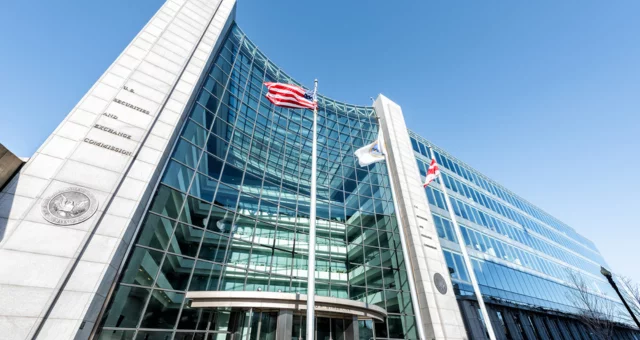 The U.S. Securities and Exchange Commission headquarters features a modern glass facade with tall, white stone columns on each side. Three flags, including the American flag, fly in front of the curved entrance under a clear blue sky.