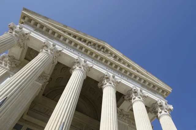 Low-angle view of a grand neoclassical building with tall Corinthian columns and ornate architectural details, set against a clear blue sky. The intricate carvings and symmetrical design convey authority and formality, often associated with government or