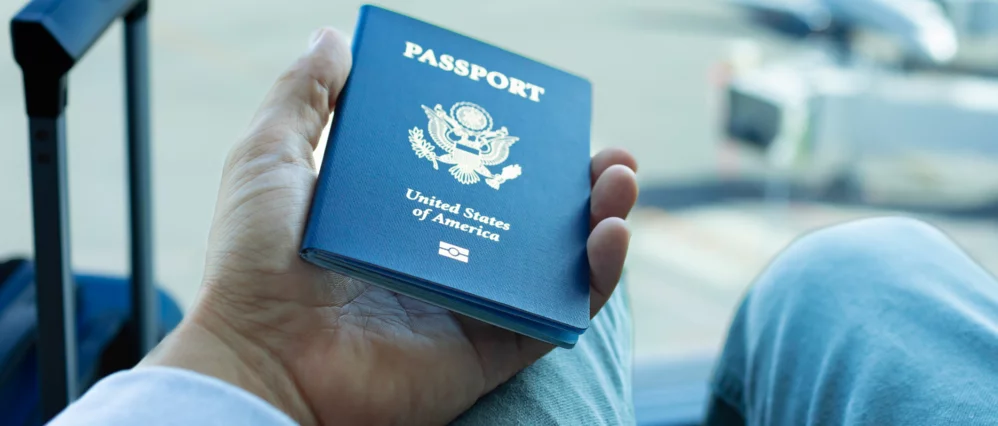 Close up of a person holding a blue United States passport with the gold emblem and the words Passport and United States of America clearly visible on the cover. The background is softly blurred, showing airport seating and luggage, suggesting travel or d