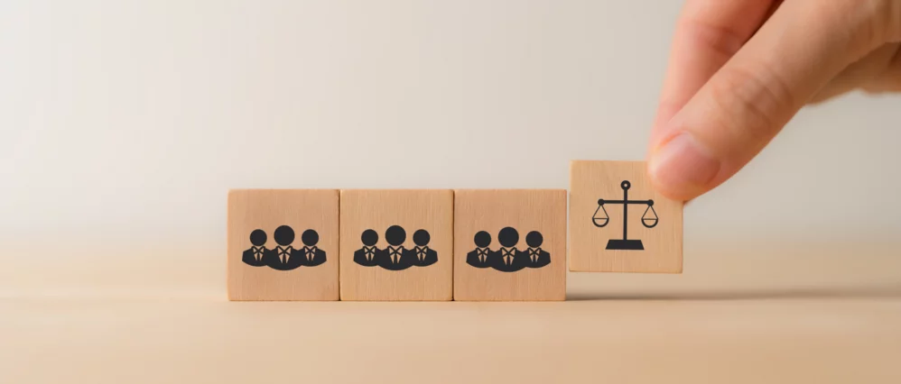 Close up of a hand placing a wooden cube with a scales of justice icon next to blocks showing groups of employees. The row of cubes features team symbols, with the justice scale block positioned at the end to emphasize legal oversight.