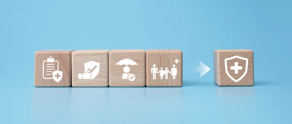 Row of wooden blocks displaying health related icons including a medical checklist, heart care, protection umbrella, and family healthcare symbols on a blue background. An arrow points toward a separate block with a shield and medical cross icon.