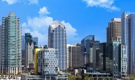 Modern city skyline featuring sleek high-rise buildings with glass facades, including a distinctive pyramid-roofed tower at the center. The scene is set under a clear blue sky with scattered clouds.