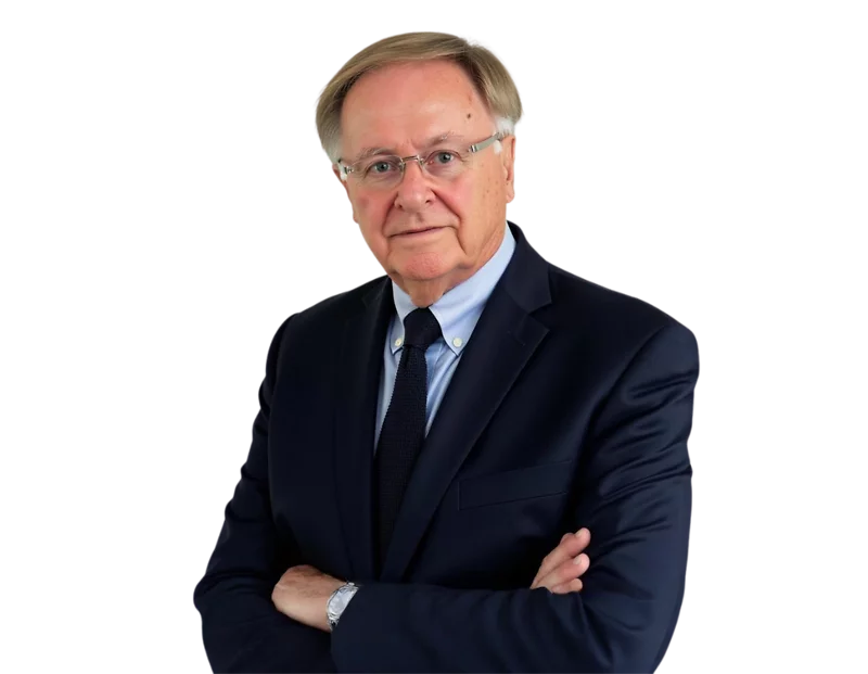Older man with glasses wearing a navy blue suit and black tie, standing with arms crossed against a white background. He has a serious expression and short, neatly combed gray hair.