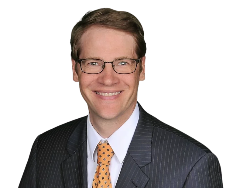 A smiling man in a dark pinstripe suit, white dress shirt, and orange patterned tie poses against a plain background. He has short light brown hair, wears rectangular glasses, and appears in a professional headshot.