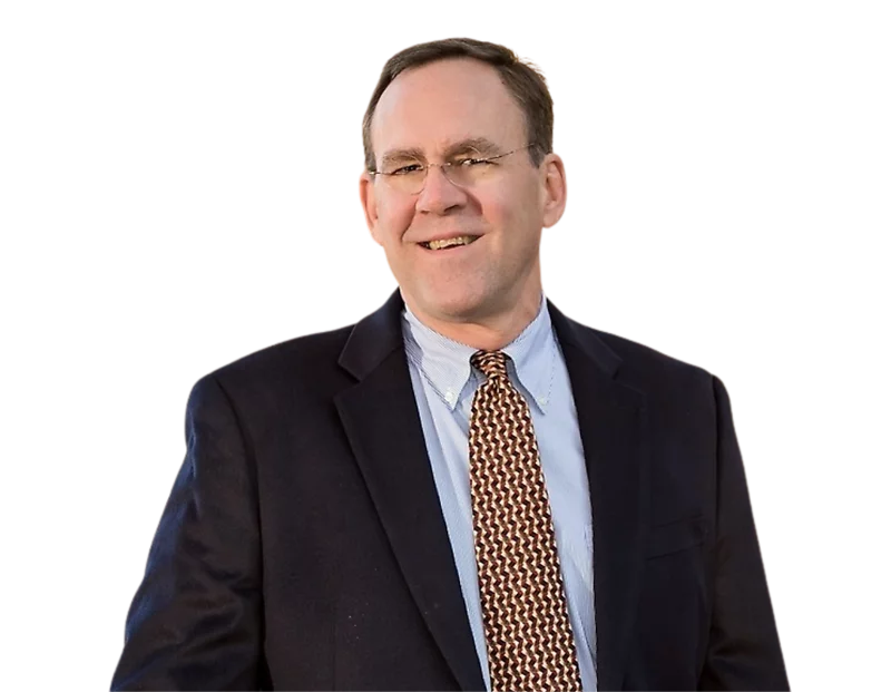 Jim Coleman in a dark blazer, light blue button-down shirt, and a geometric patterned tie, standing confidently against a transparent background. His relaxed posture and business attire convey a professional yet approachable demeanor.