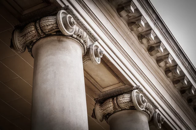 Close-up view of classical architectural columns with ornate Ionic capitals, featuring scroll-like volutes and carved detailing. The weathered stone and perspective highlight the elegance and strength of historic building design.