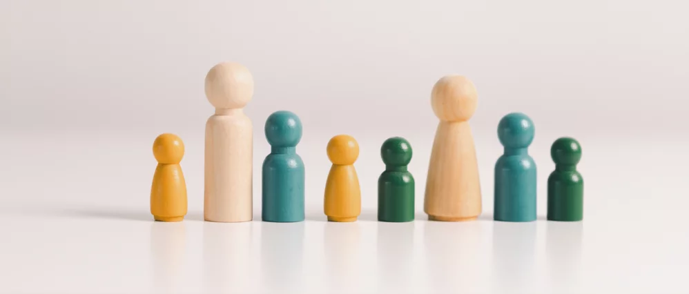 Row of wooden peg figures in different sizes and colors arranged side by side on a neutral background. Two taller natural wood figures stand among shorter yellow and green figures, suggesting variation in roles or hierarchy within a group.