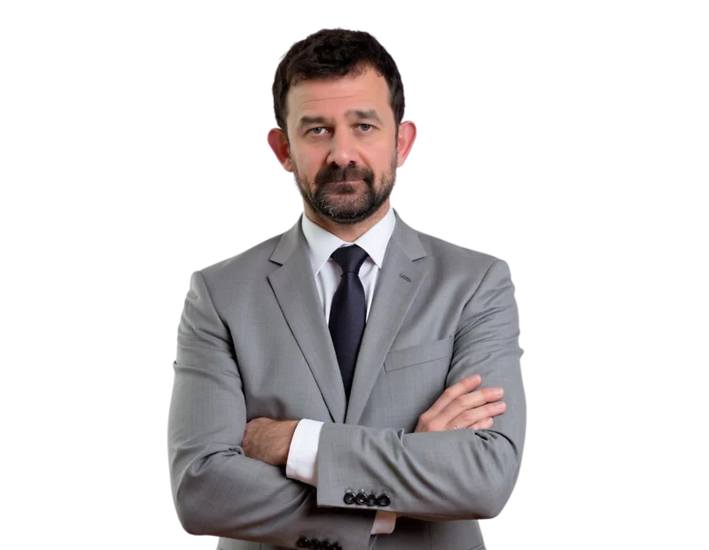 Serious-looking man with short dark hair and beard wearing a light gray suit and dark tie, standing with arms crossed against a transparent background. His formal attire and posture suggest a professional or executive portrait.