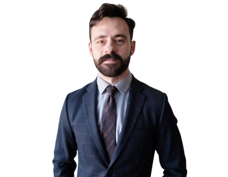 Man with short brown hair and a beard wearing a dark blue checkered suit, light blue dress shirt, and patterned tie, standing against a black background.