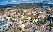 Aerial view of downtown Asheville, North Carolina, with a mix of historic brick buildings and modern mid-rises. The city is surrounded by green hills and mountains under a partly cloudy sky.