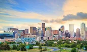 A panoramic view of Denver’s downtown skyline at sunset, with modern high-rise buildings reflecting soft golden light. In the foreground, a curved highway and green park space lead toward the city, while the blue arch of a bridge stands out against the ur