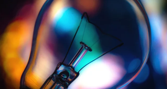 Close-up photograph of a clear light bulb against a blurred background of vivid blue, orange, and purple bokeh lights. The bulb’s delicate filament and glass structure are sharply in focus, contrasting with the soft, colorful glow behind it.