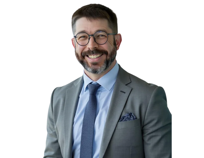 Smiling professional man with short dark hair, beard, and round glasses, wearing a light gray suit with a light blue shirt, patterned pocket square, and navy tie. He stands against a transparent background, giving off a warm and approachable executive pre