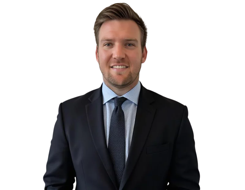 Jadie Mims in a dark suit with a light blue dress shirt and navy patterned tie is smiling at the camera. He has short, neatly styled brown hair and light facial stubble.