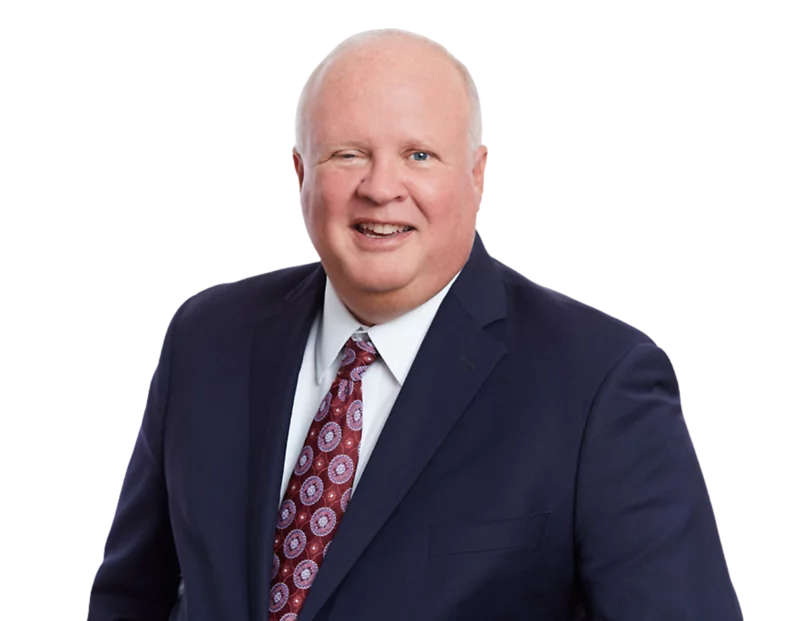 Smiling older man in a dark navy suit with a white shirt and maroon tie featuring a circular pattern, posed against a transparent background. His formal outfit and approachable demeanor suggest a senior professional or executive portrait.