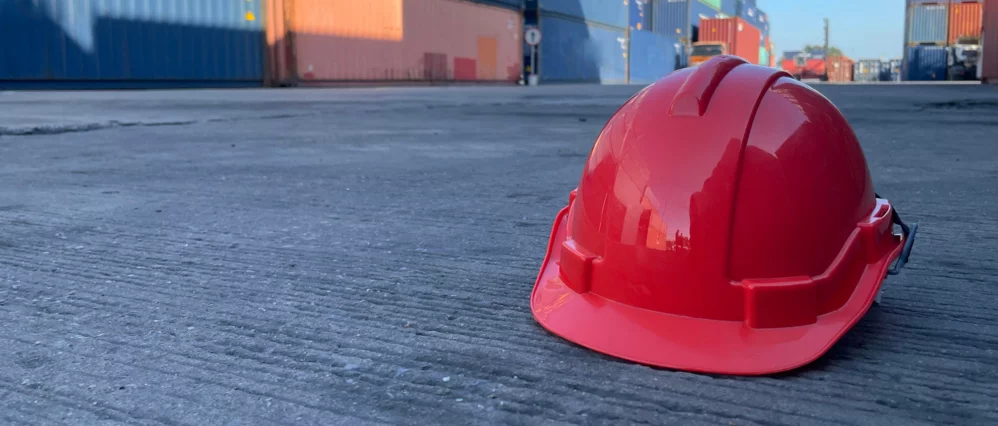 Close up of a red safety hard hat resting on a concrete surface at an industrial site. Shipping containers and equipment appear blurred in the background, suggesting a construction or logistics environment.