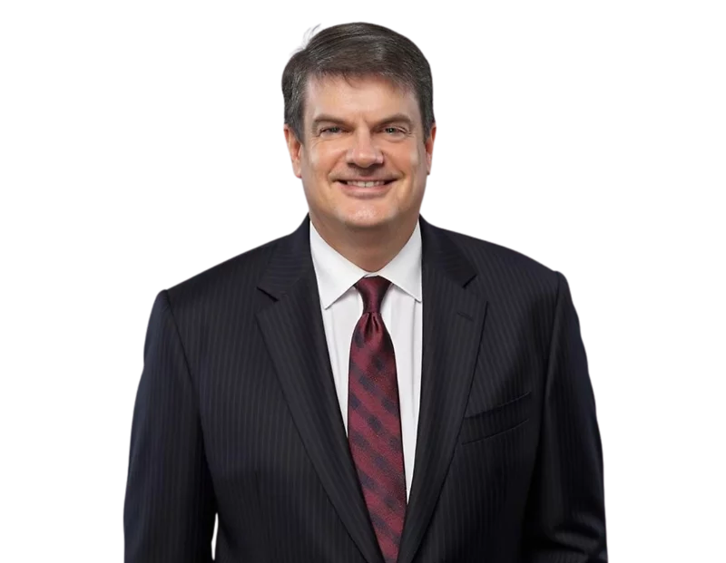 A professional headshot of a man wearing a dark pinstripe suit, white dress shirt, and a maroon patterned tie, smiling confidently. The background has been removed, making the image suitable for corporate profiles or executive bios.