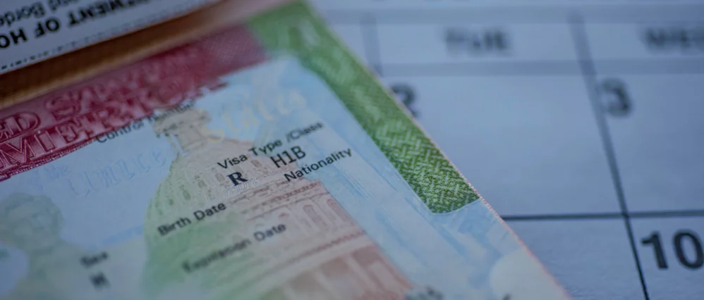 Close up of an H1B visa stamp inside a passport with detailed text and security patterns visible. A blurred calendar page appears in the background, suggesting a specific date or deadline.
