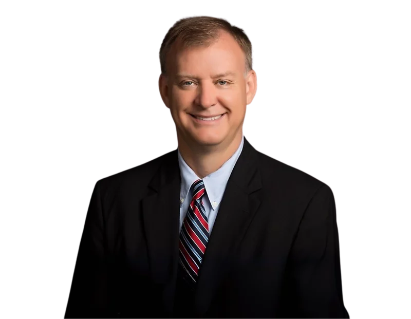 Man with short light brown hair smiling at the camera, wearing a black suit jacket, light blue shirt, and a red, white, and blue striped tie, posed against a black background.