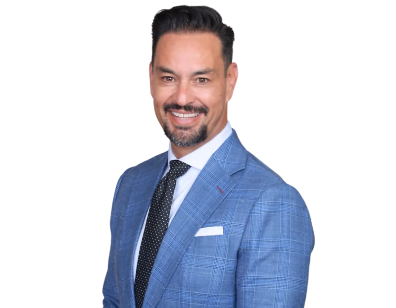 Guillermo Escobedo in a light blue plaid suit jacket, white shirt, and black polka-dot tie smiles against a light blue and white geometric background.