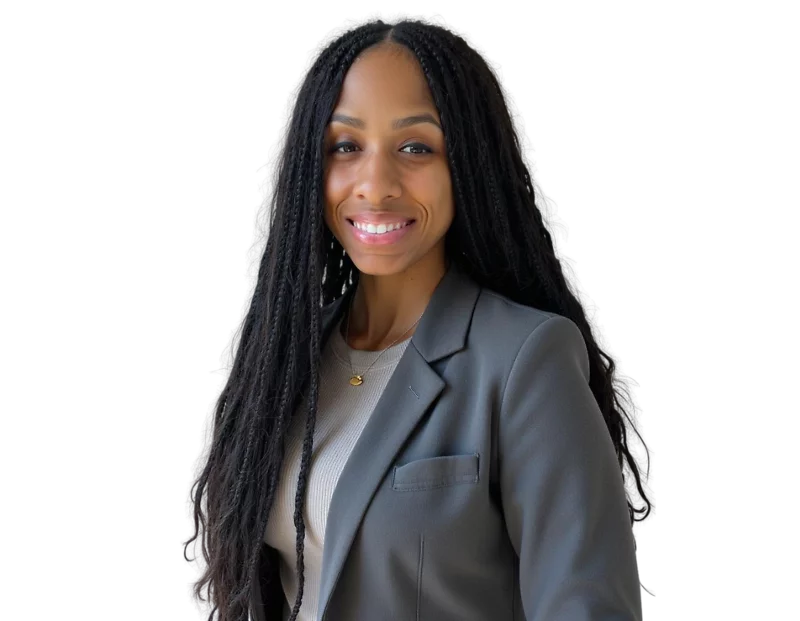 A photo of Nia Doaks smiles confidently while wearing a gray blazer over a light beige top. She stands against a transparent background, dressed in professional attire with a gold pendant necklace, exuding warmth and professionalism.