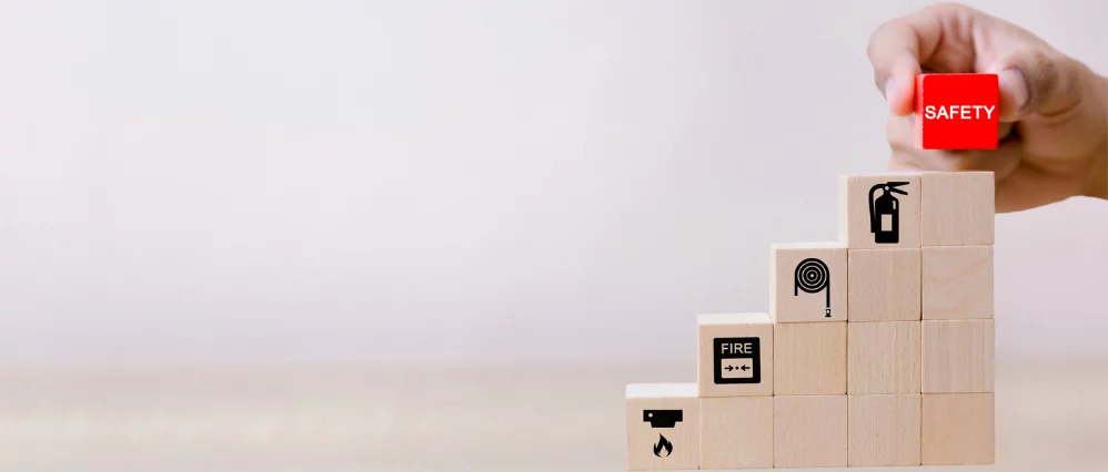 Close up of a hand placing a red cube labeled SAFETY on top of stacked wooden blocks arranged like steps. The lower blocks display fire safety icons including a flame, fire alarm, hose reel, and fire extinguisher.